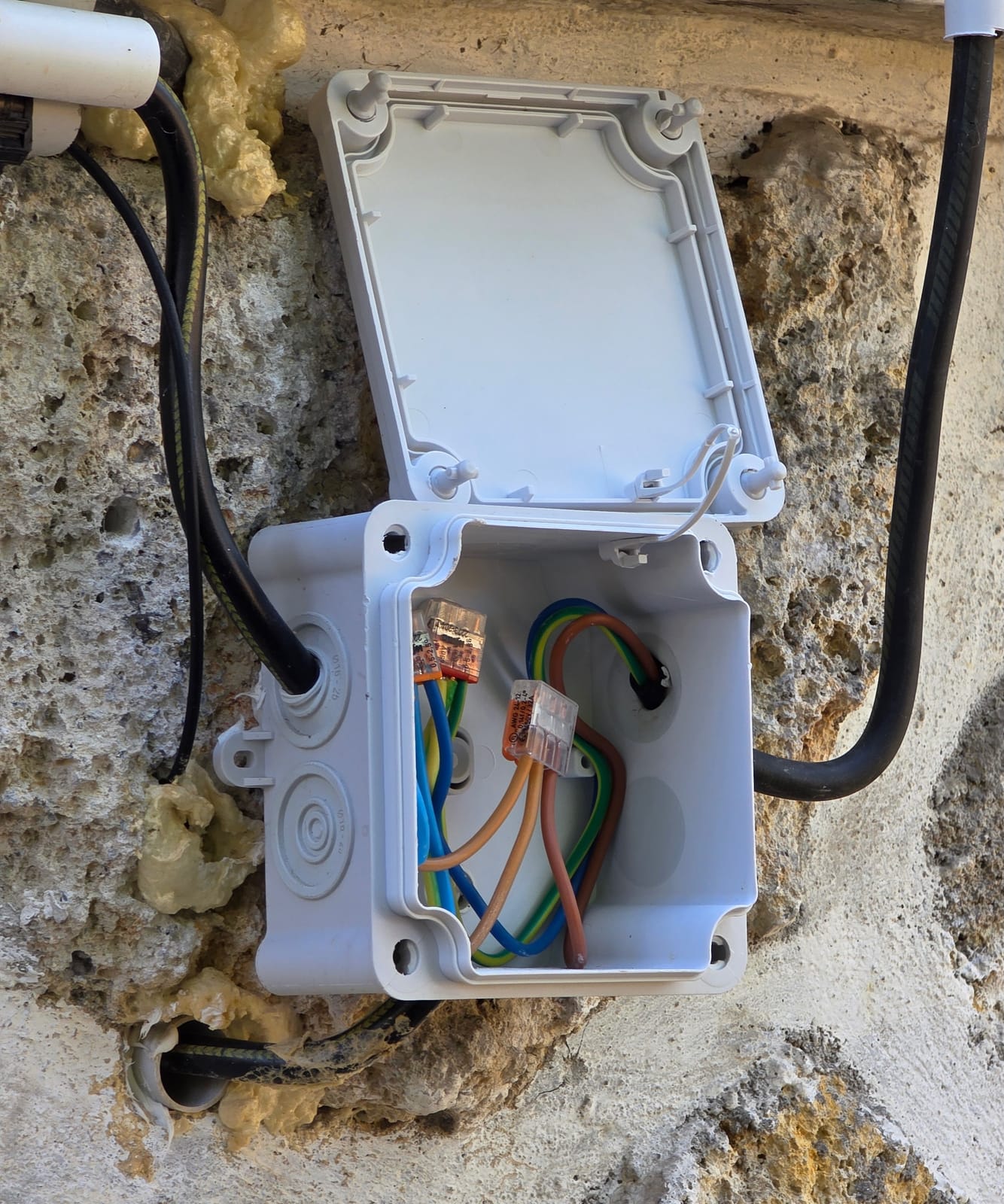 Boîte de dérivation extérieure ouverte sur un mur en pierre avec câbles, connecteurs et mousse expansive