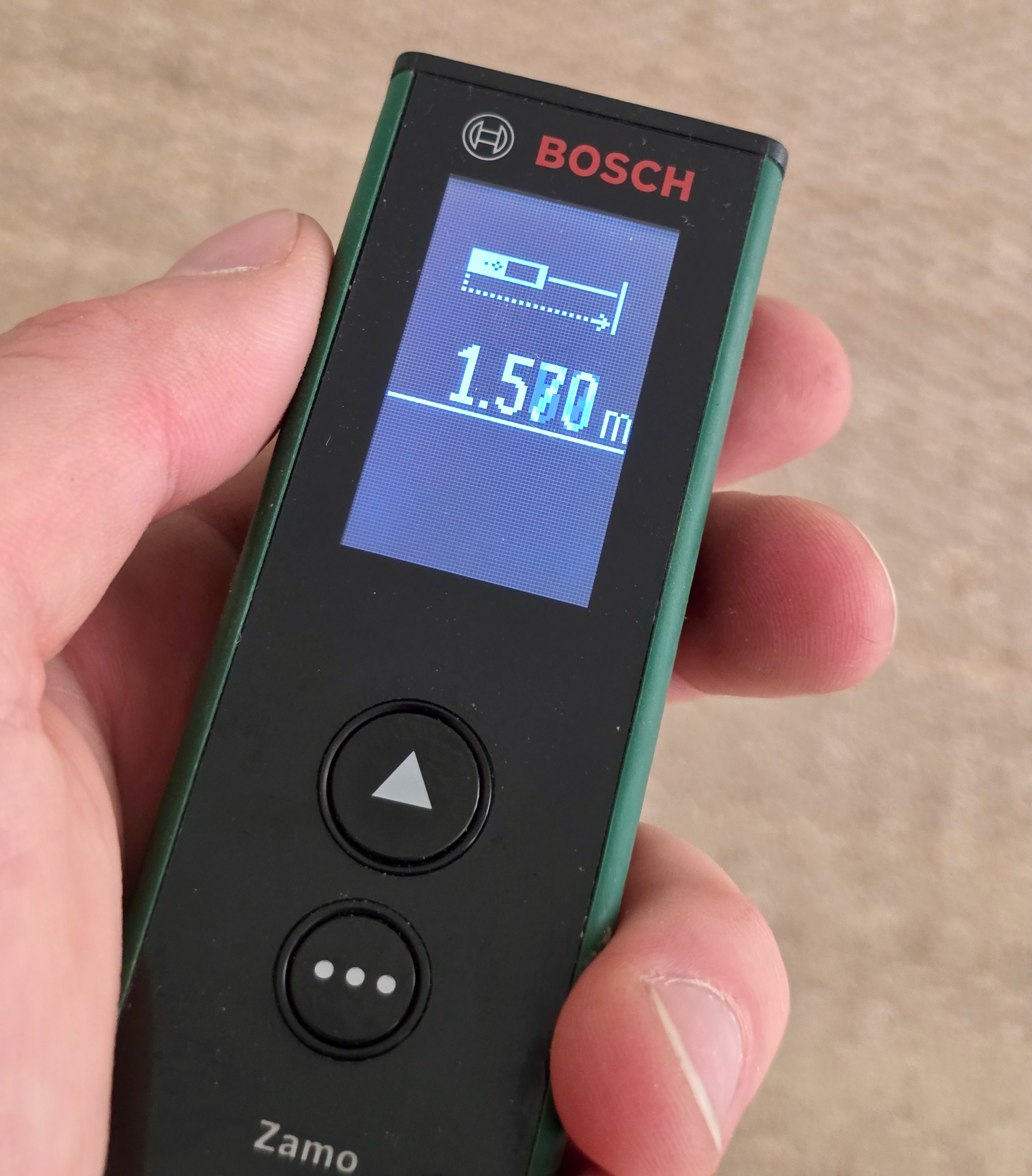 Télémètre laser Bosch Zamo tenu en main, écran affichant une mesure de distance et deux touches rondes en dessous.