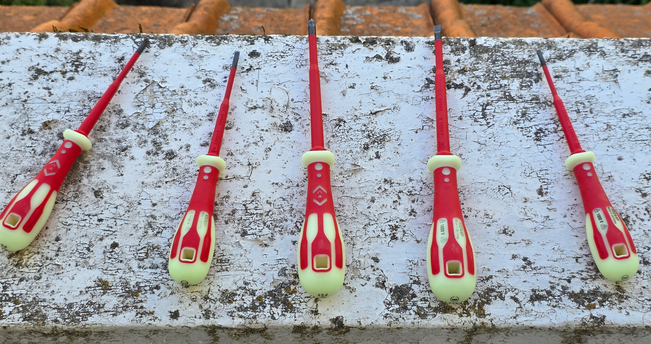 Fünf C.K Tools VDE-Schraubendreher mit roten und phosphoreszierenden Griffen, aufgereiht auf einer kleinen weißen Mauer im Freien