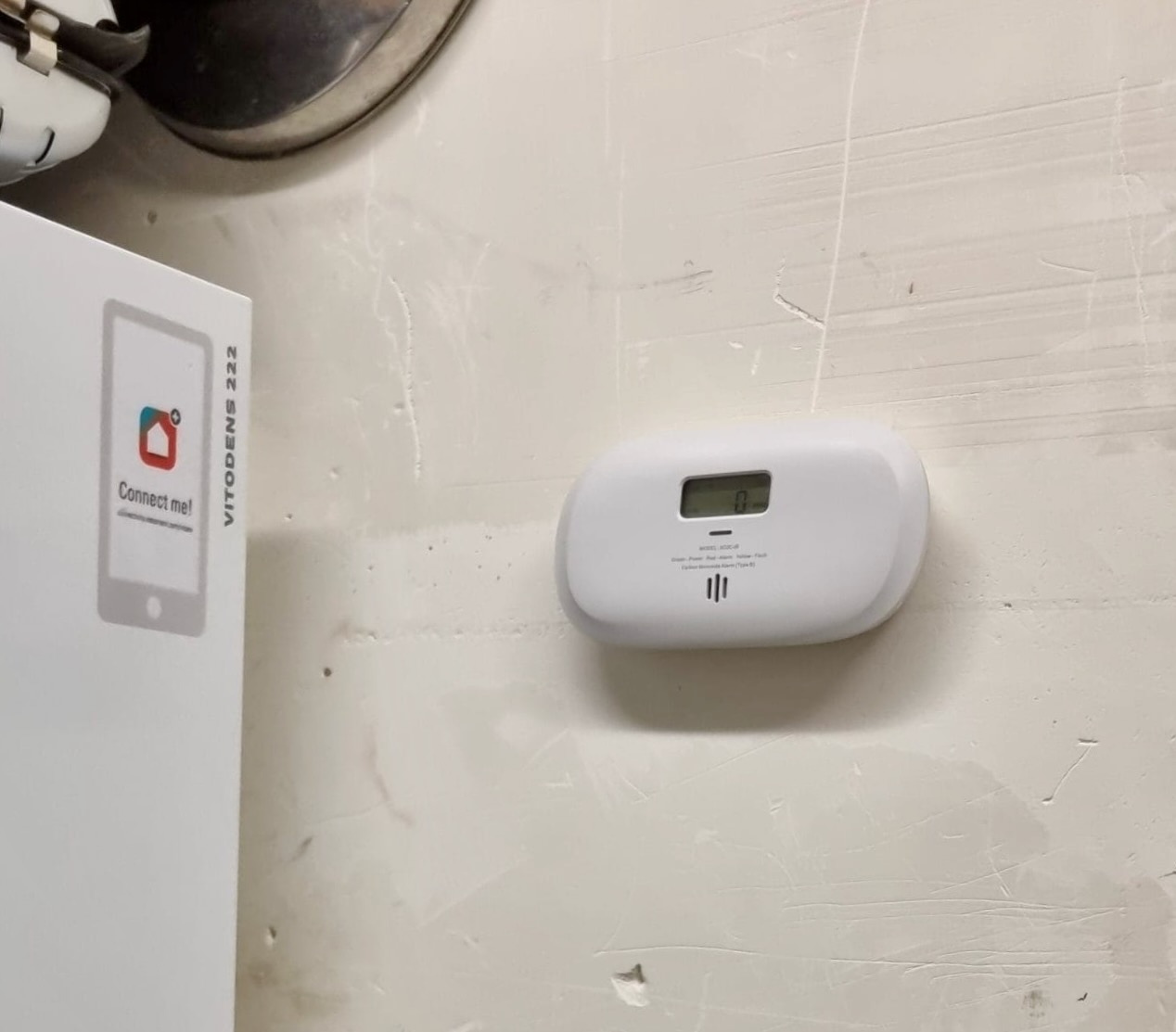 Détecteur de monoxyde de carbone X-Sense fixé au mur, écran LCD affichant 0 ppm, à côté de la chaudière blanche