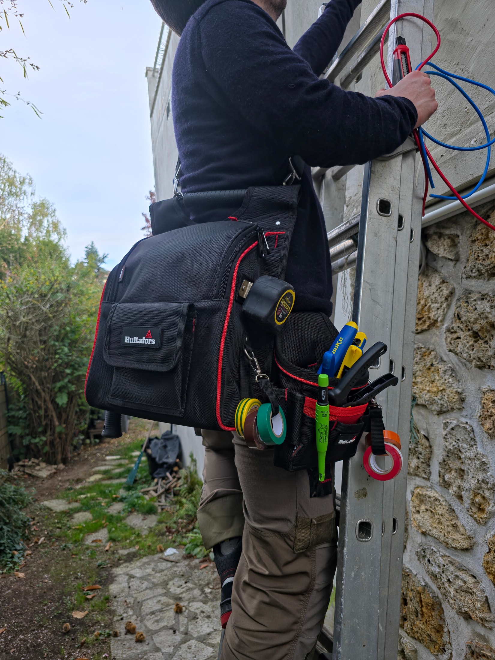 Électricien sur une échelle aluminium avec une sacoche Hultafors à la hanche, câbles bleus et rouges en main