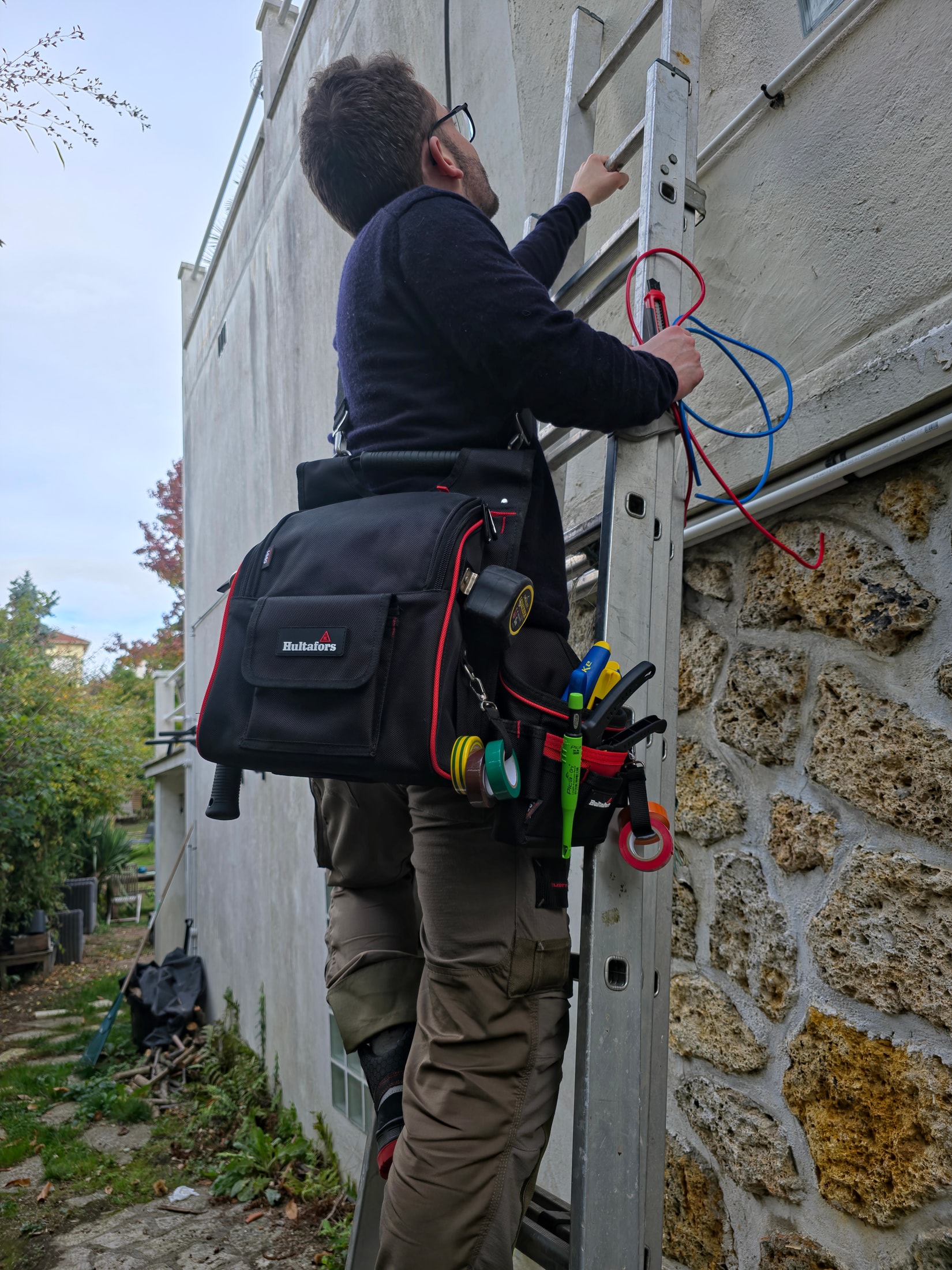 Électricien sur une échelle aluminium avec une sacoche Hultafors en bandoulière, travaillant sur une façade