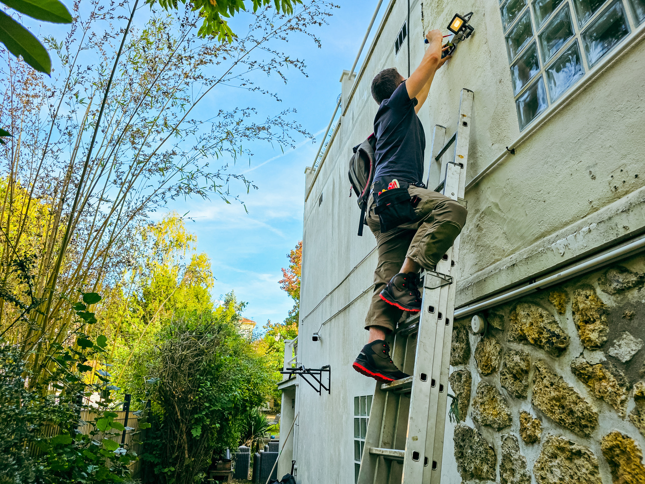 Électricien sur une échelle en aluminium installant un projecteur mural sur une façade blanche
