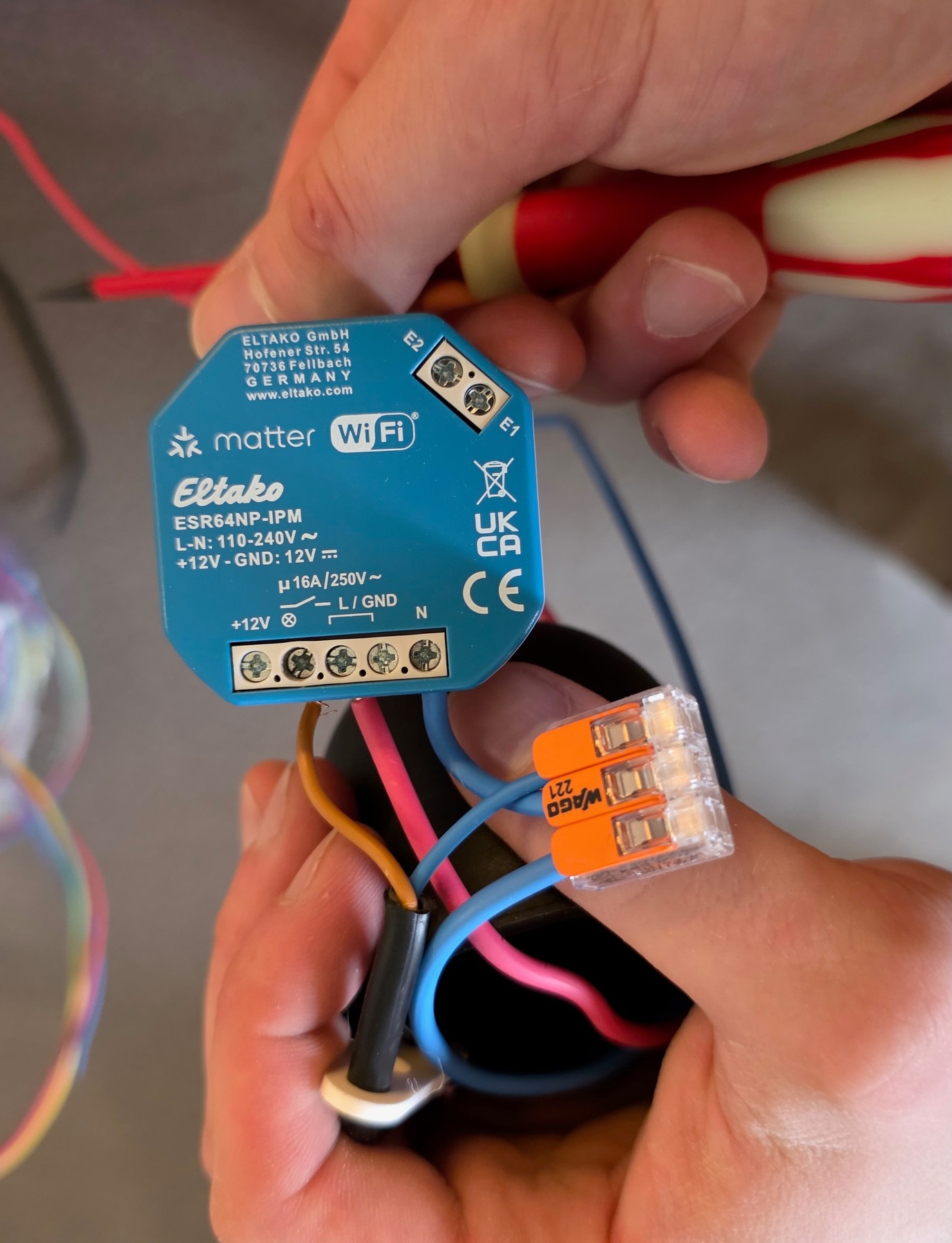 Mains en train d’équiper un micromodule Eltako ESR64NP-IPM au-dessus d’un pavillon de plafond noir, tournevis en main et conducteurs reliés par un connecteur Wago.