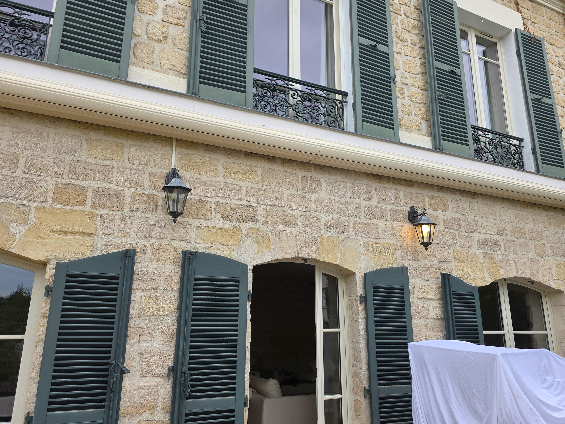 Façade en pierre, volets verts et deux lanternes murales, bâche blanche au premier plan