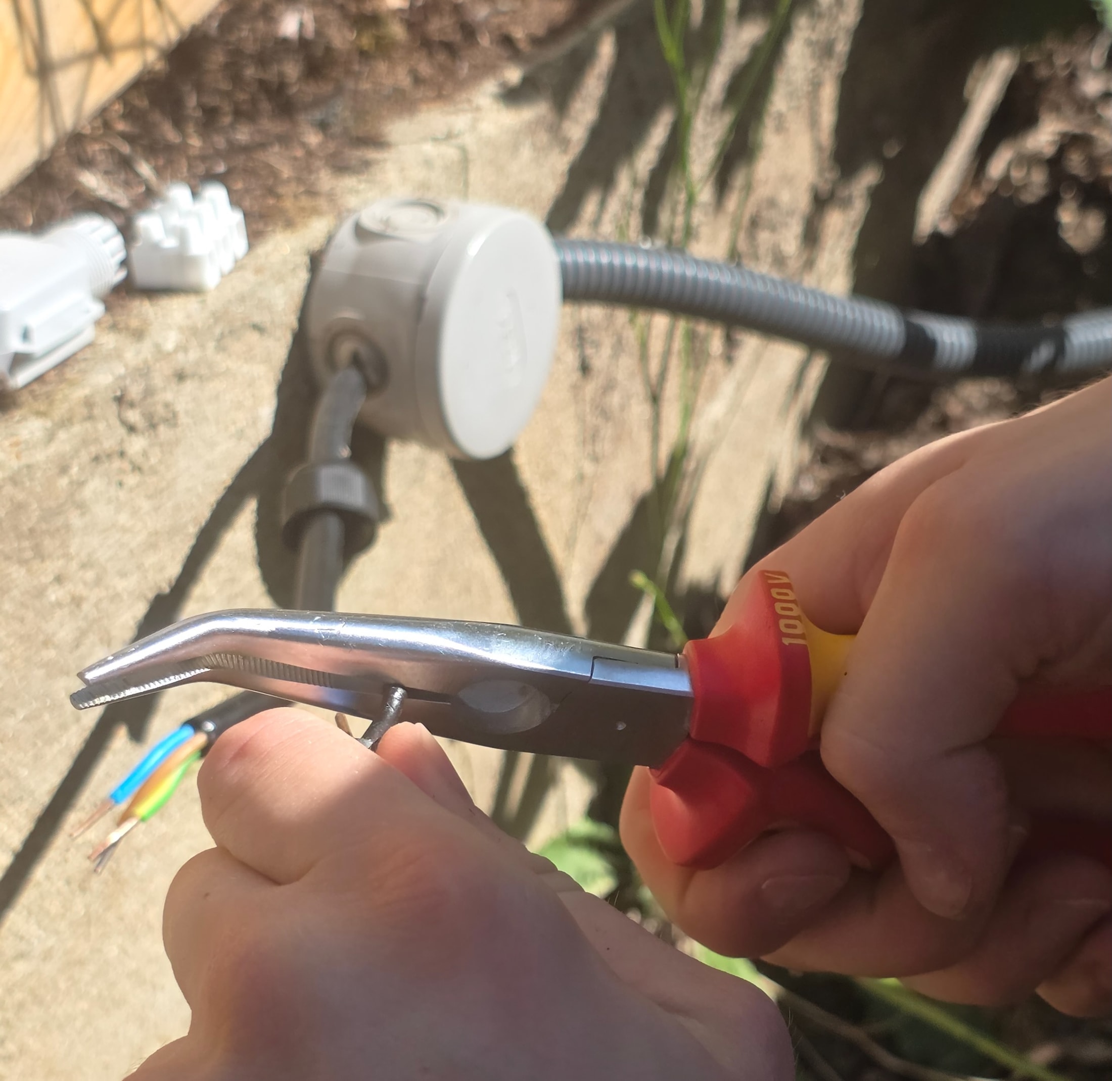 Main tenant un fil avec une pince à bec coudée isolée 1000 V devant une boîte de dérivation, pour réparer un fil de phase brûlé par un ancien connecteur Amazon
