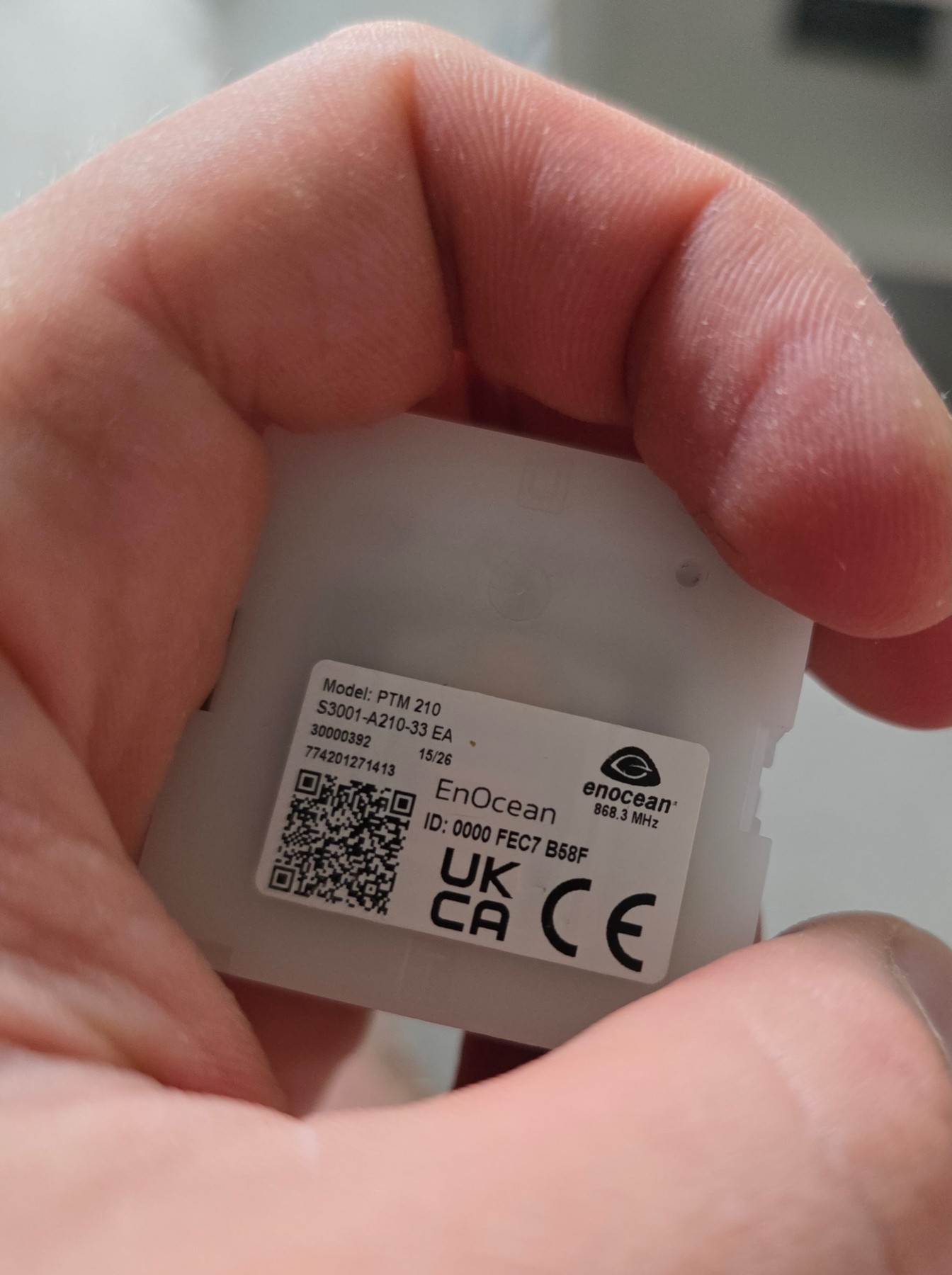 Dos d’un module radio EnOcean PTM 210 tenu en main, avec une étiquette portant le modèle, l’identifiant radio et la fréquence 868,3 MHz.