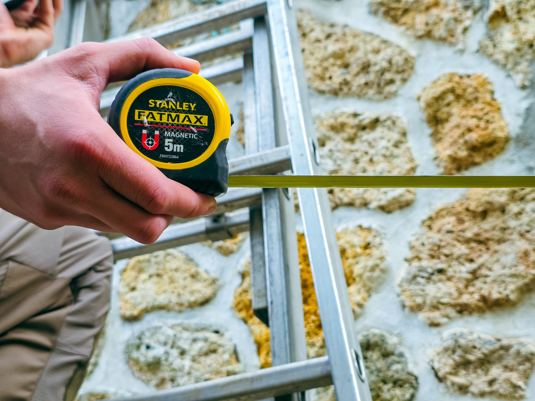 Mètre ruban Stanley FatMax Magnetic 5 m tenu en main devant un mur en pierre et une échelle