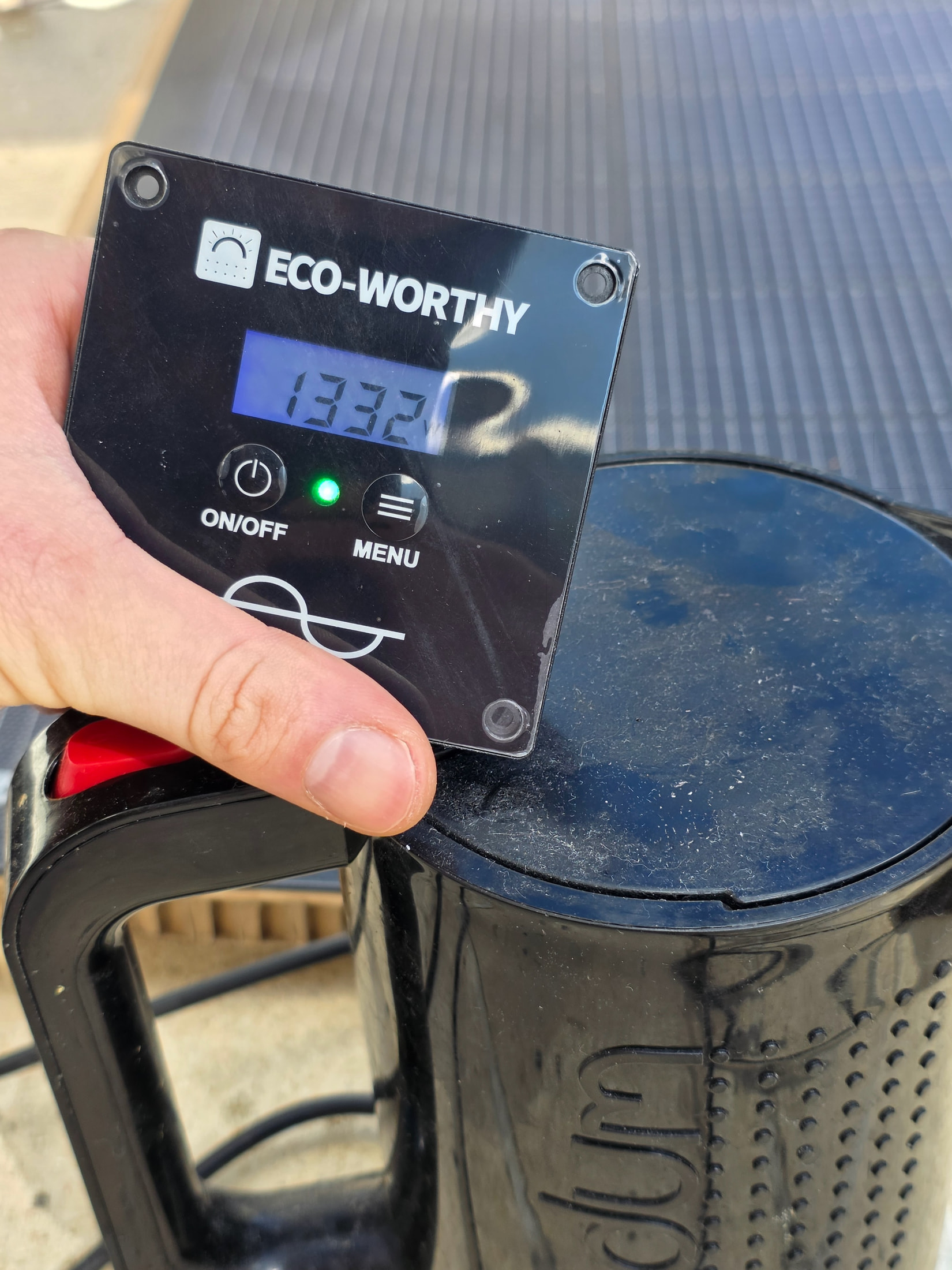 Afficheur de l’onduleur ECO-WORTHY indiquant 1332 W, tenu à la main devant la bouilloire noire