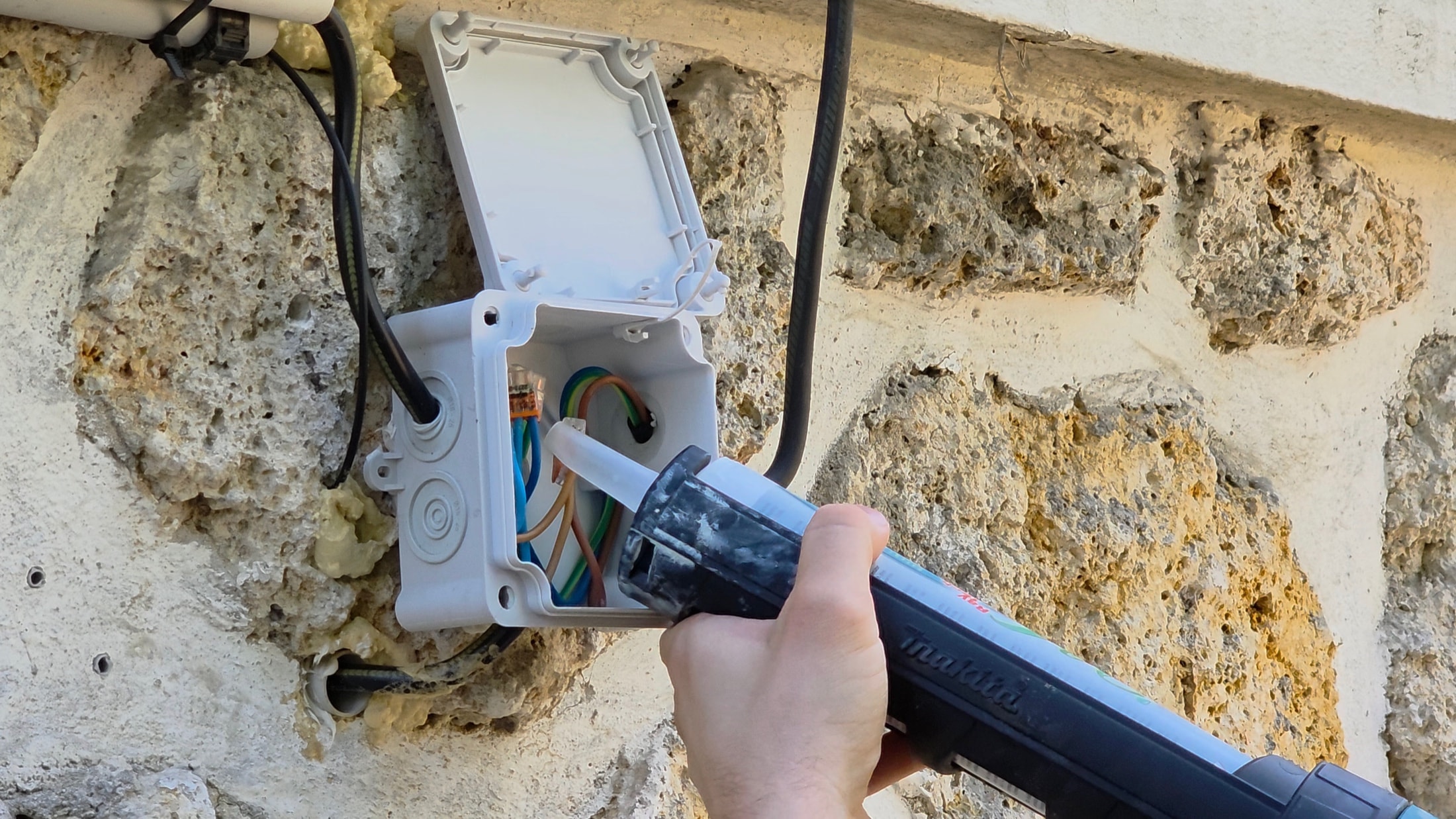Pistolet à mastic Makita appliquant le gel isolant dans une boîte de dérivation sur un mur en pierre