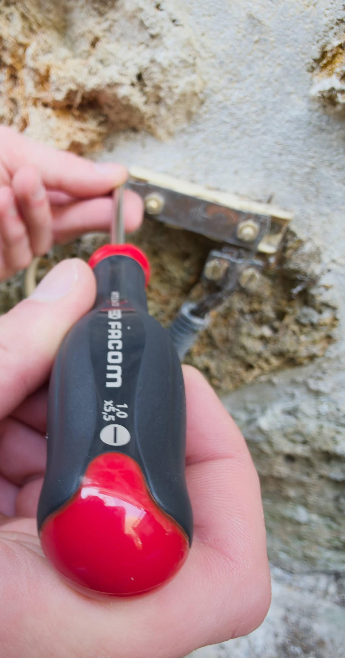 Tournevis FACOM AT5.5X125 tenu devant une barrette de terre fixée sur un mur en pierre