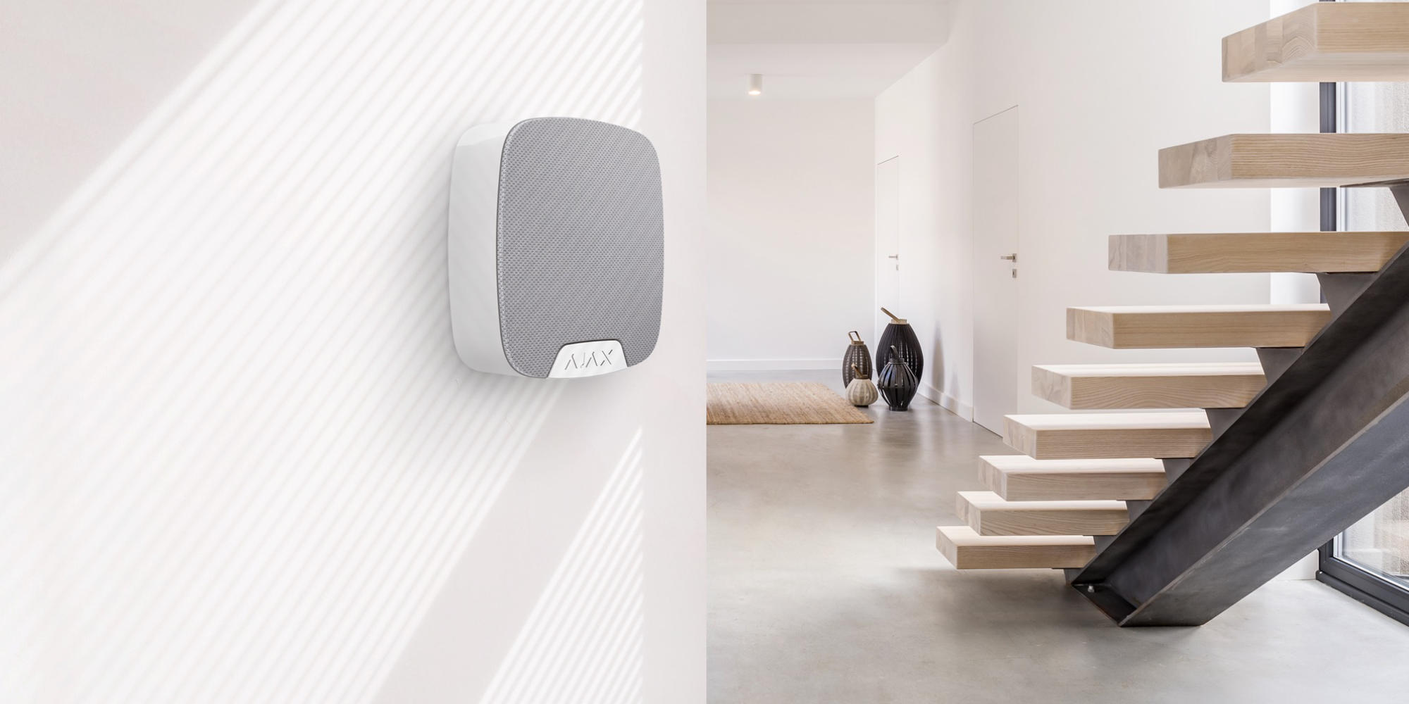 Une sirène Ajax blanche fixée sur un mur blanc dans un intérieur moderne avec escalier en bois.