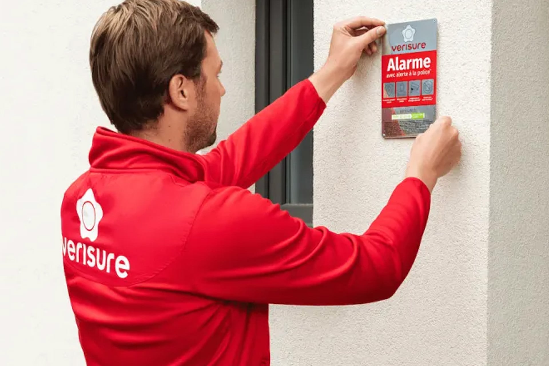Technicien portant un sweat rouge à l’effigie de Verisure, en train d’installer un panneau d’alarme sur un mur extérieur.