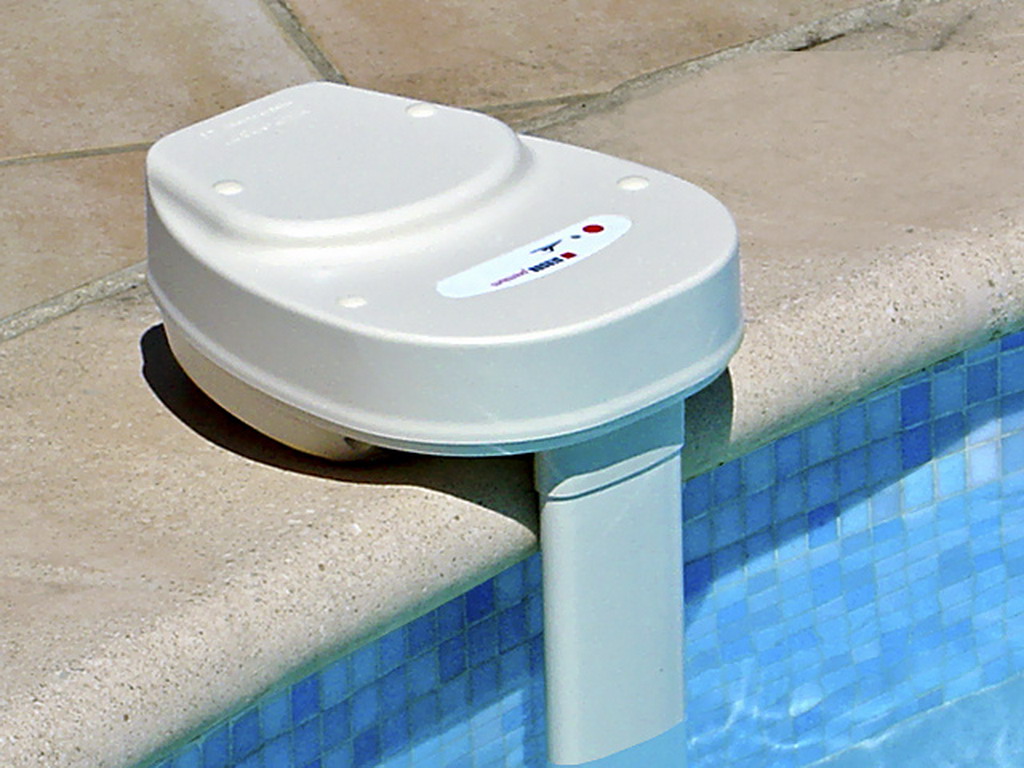 Un boîtier blanc d'alarme fixé sur le rebord d'une piscine carrelée avec une sonde descendant dans l'eau.