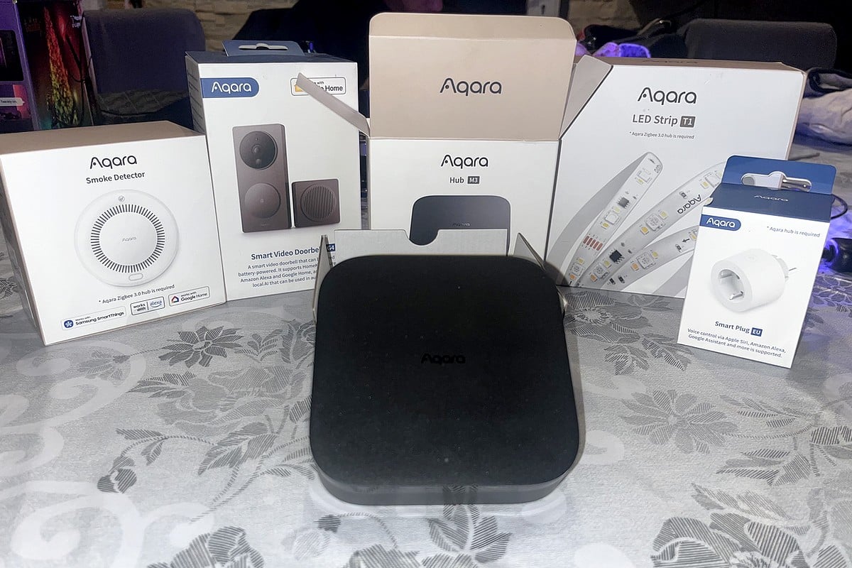 Ensemble de produits et boites Aqara disposés sur une table, incluant un détecteur de fumée, une sonnette vidéo, un hub M3 noir, une bande LED et une prise connectée.
