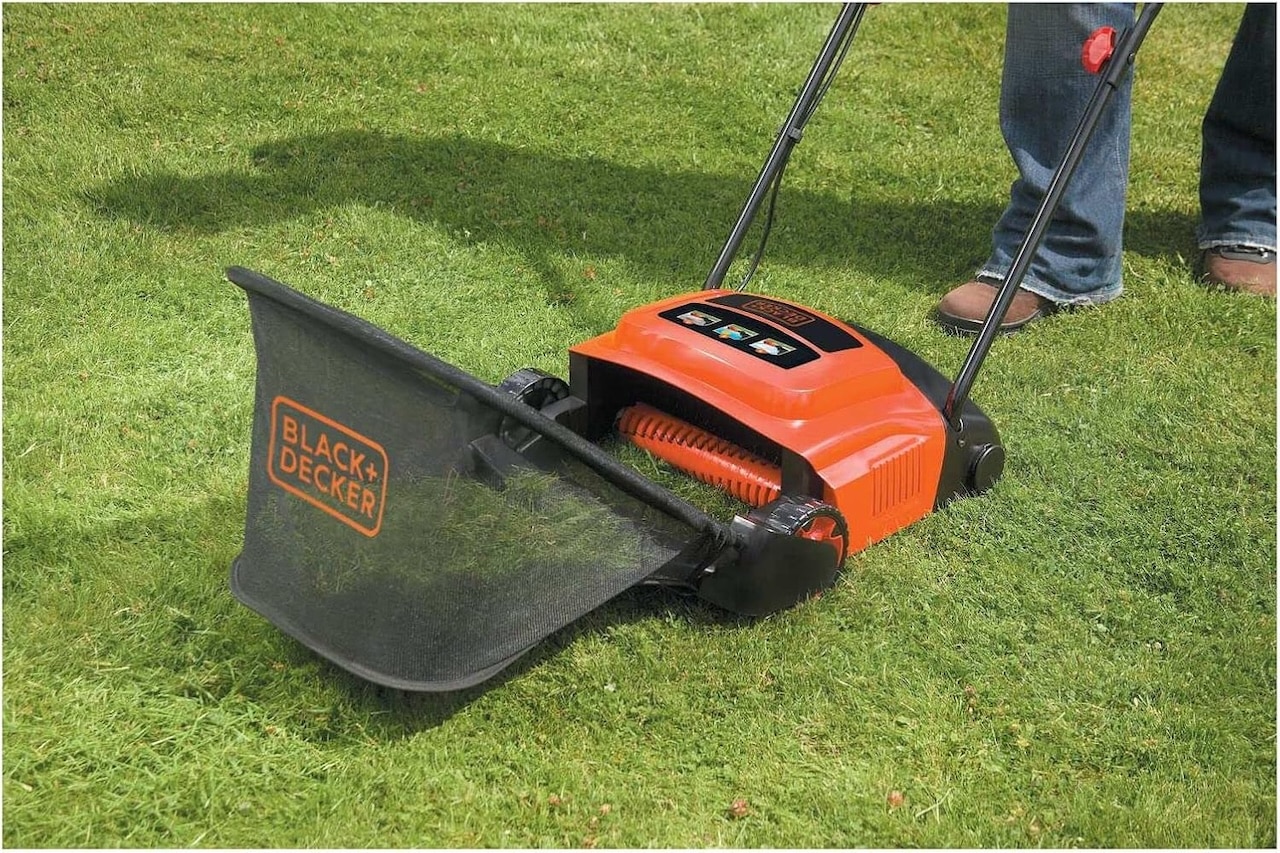 Un scarificateur de gazon orange et noir Black+Decker avec sac de ramassage est poussé sur une pelouse verte.