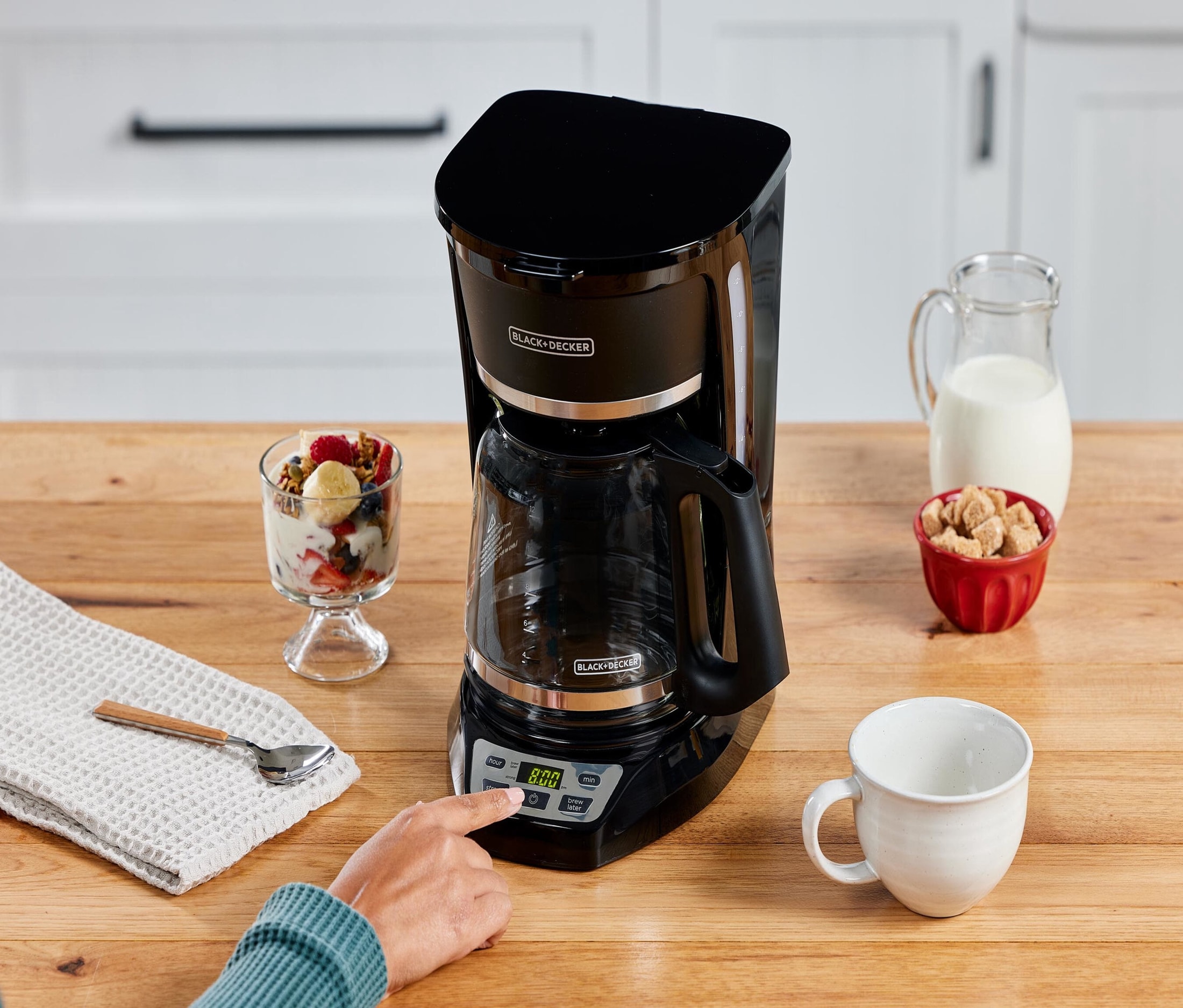 Une cafetière filtre noire Black+Decker posée sur un plan de travail en bois avec une tasse et des aliments autour.