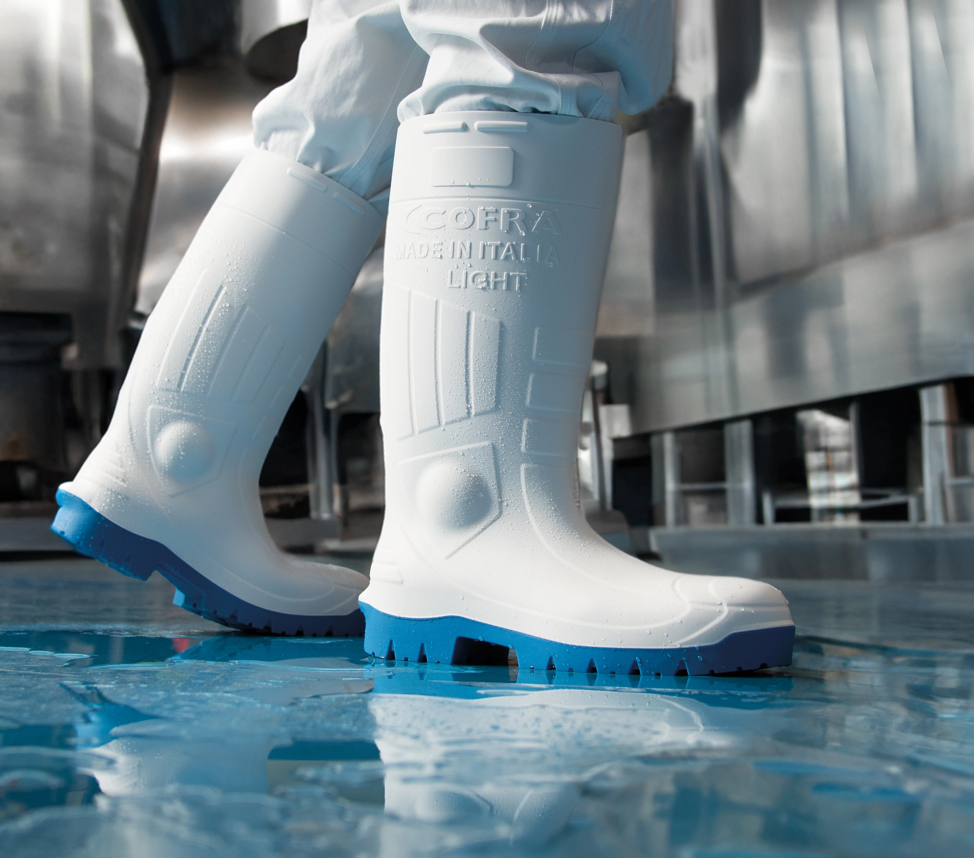 Bottes blanches en caoutchouc à semelle bleue portées par une personne en tenue blanche dans un environnement industriel humide, inscription COFRA MADE IN ITALIA LIGHT visible.
