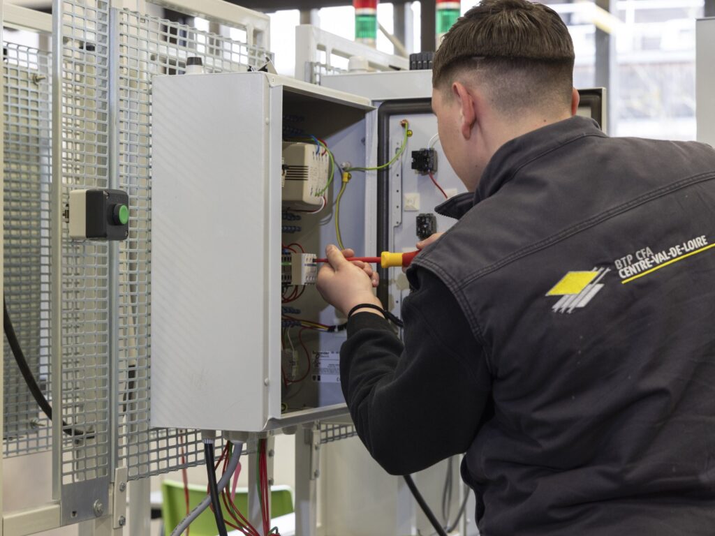 Un apprenti électricien de dos portant une veste 'BTP CFA CENTRE-VAL-DE-LOIRE' travaillant sur un tableau électrique ouvert