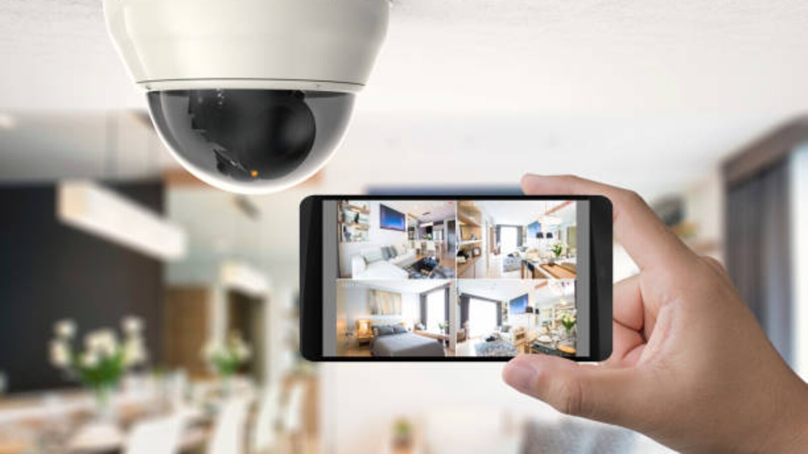 Une caméra dôme fixée au plafond se trouve au-dessus d’une main tenant un smartphone qui affiche quatre flux de surveillance de pièces intérieures.