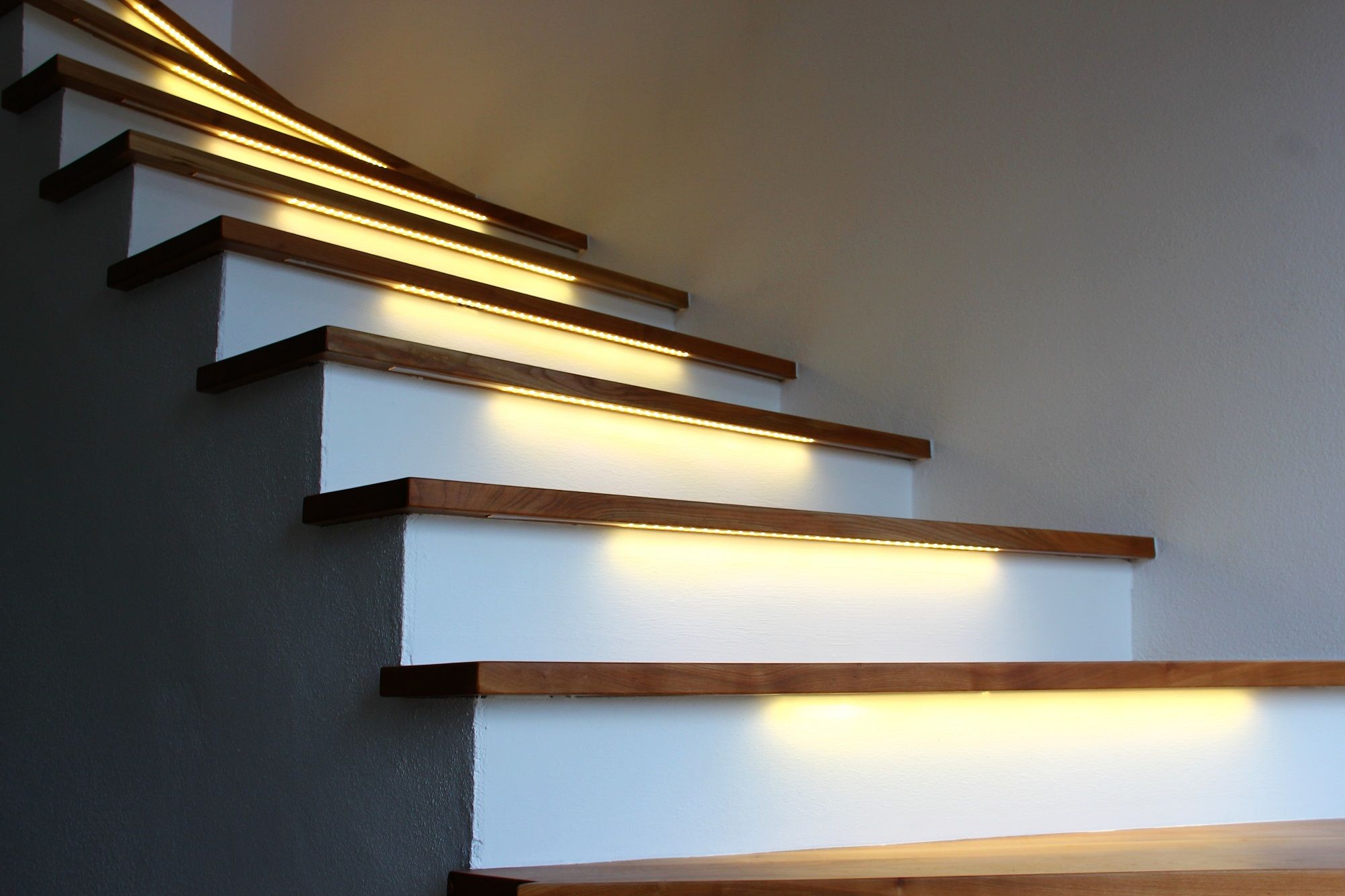 Escalier avec marches en bois et contremarches blanches, éclairé par des bandes LED installées sous chaque marche produisant une lumière chaude
