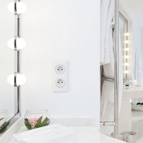 Photo d’une salle de bain blanche avec une prise double au centre, un miroir éclairé à gauche et un porte-serviettes à droite