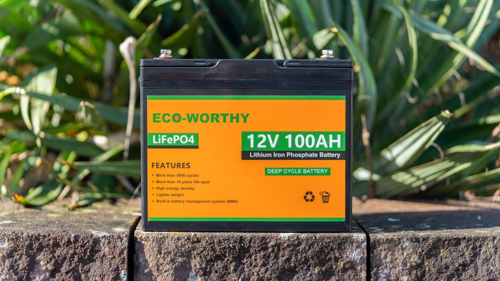 Une batterie ECO-WORTHY LiFePO4 12V 100AH posée sur un muret en pierre devant de la végétation.