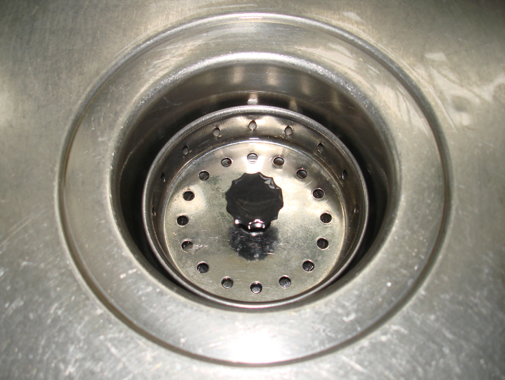 Trou d’évacuation d’un évier en inox vu de dessus, avec sa grille et sa poignée noire
