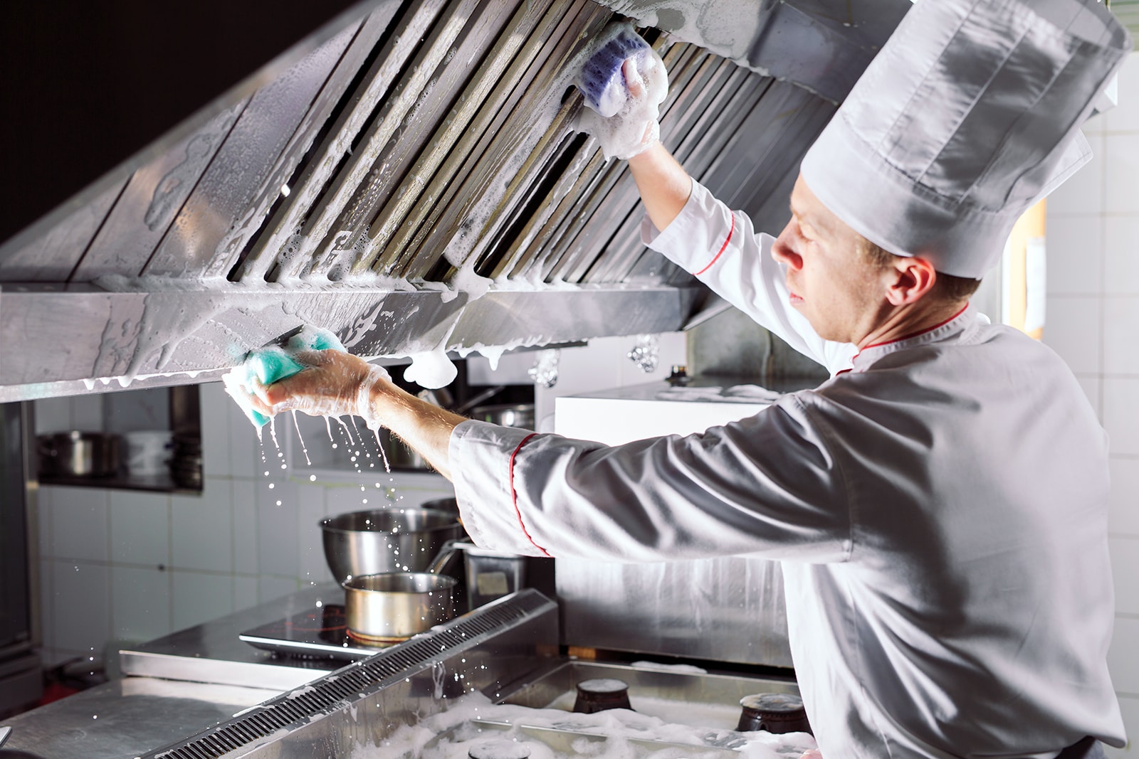 Un cuisinier en uniforme blanc et toque frotte avec une éponge savonneuse les filtres d’une grande hotte de cuisine professionnelle.