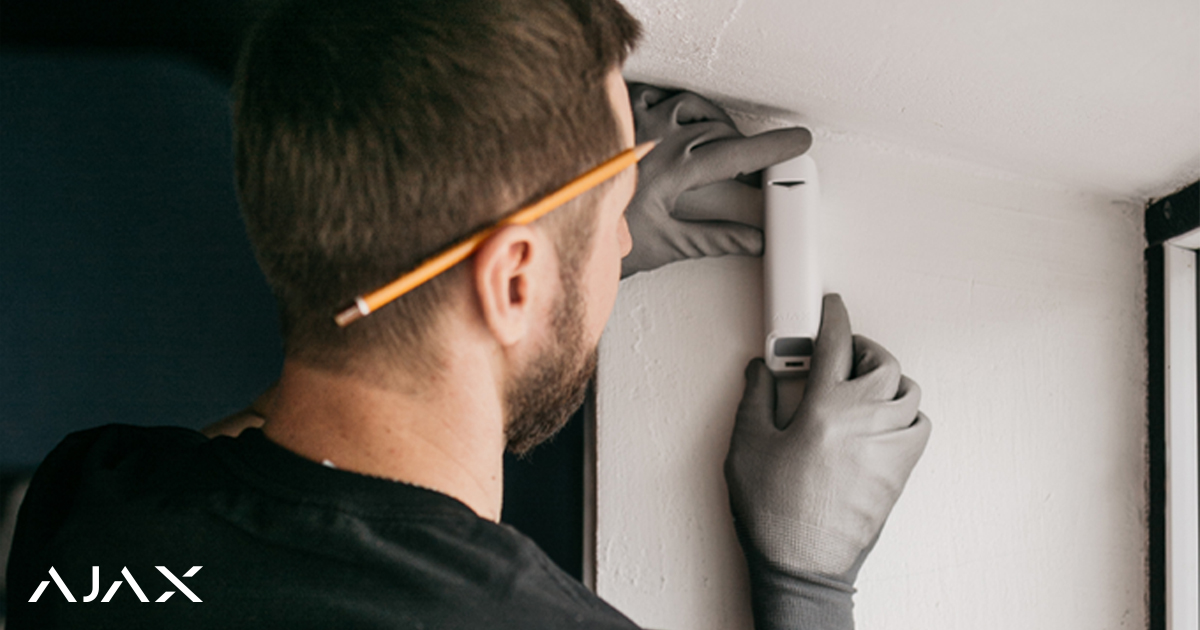Un technicien en gants installant un détecteur blanc sur un mur avec le logo Ajax en filigrane.