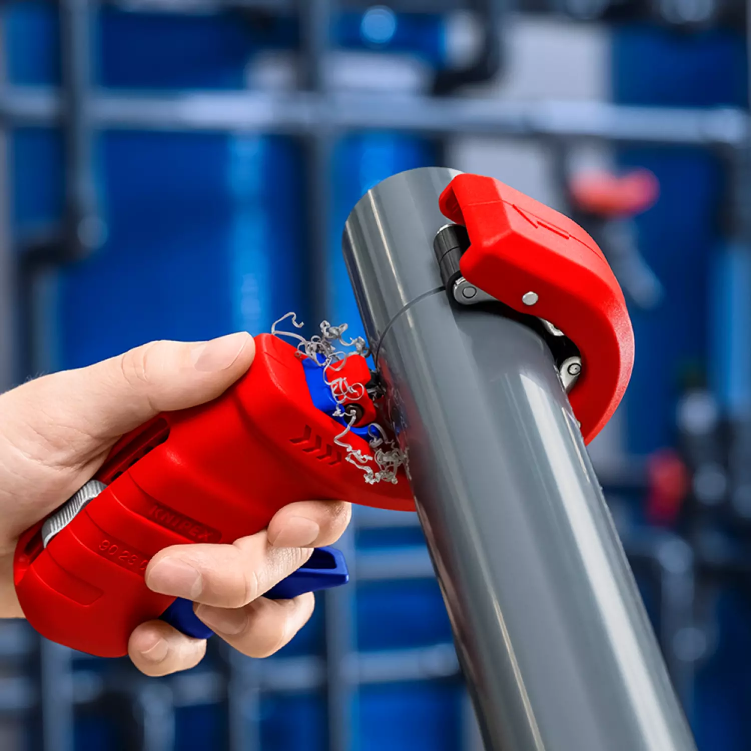 Une main utilisant un coupe-tube rouge et bleu pour sectionner un tube gris, avec des copeaux de matière visibles.
