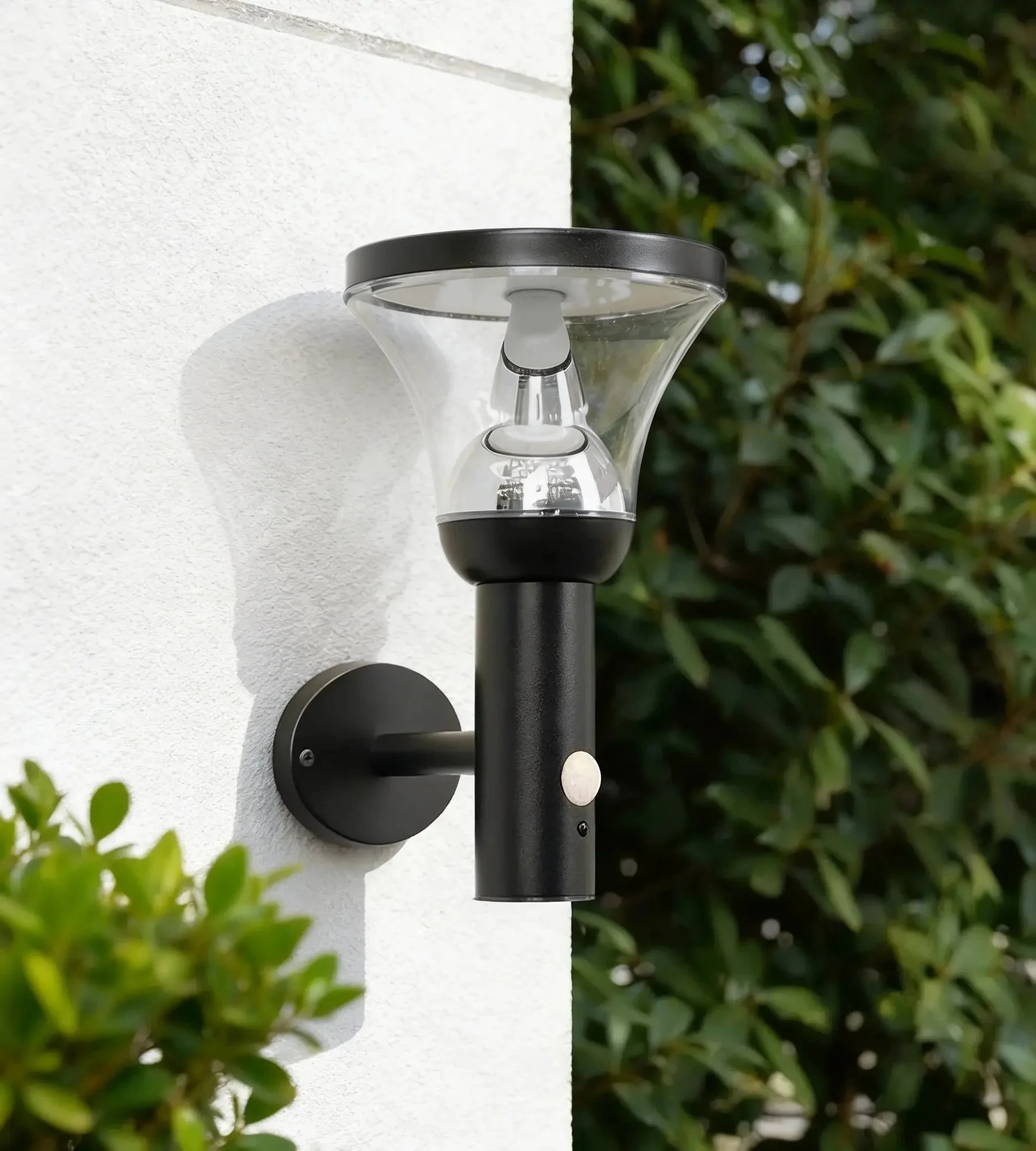 Une applique murale extérieure noire avec un globe en verre transparent et un capteur blanc sur le corps, fixée sur un mur blanc avec de la végétation en arrière-plan.