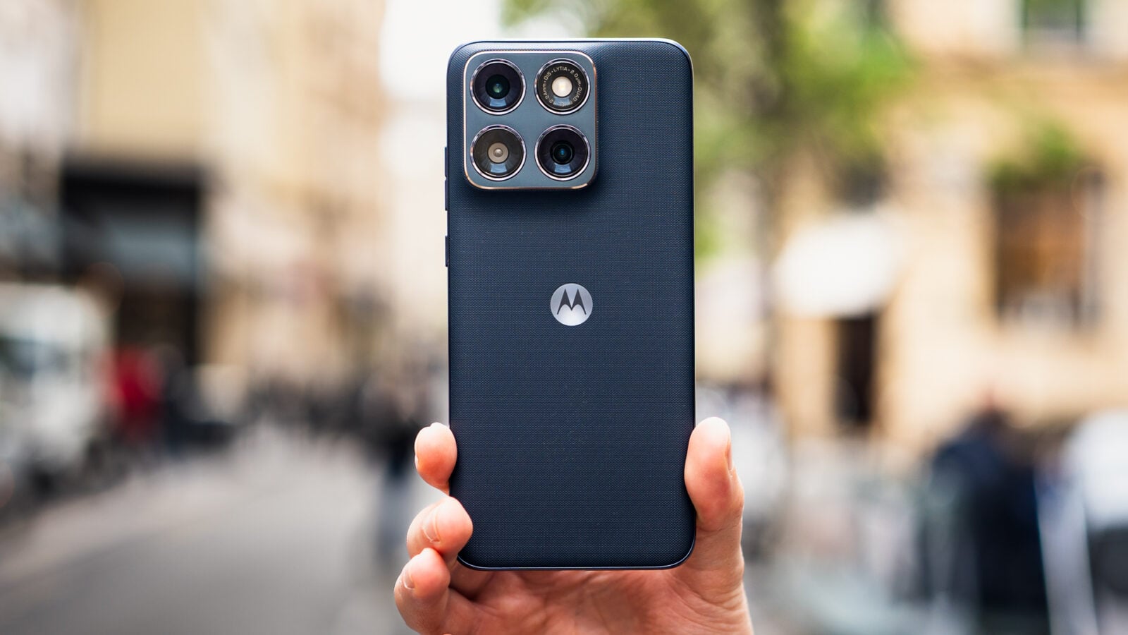 Main tenant un smartphone Motorola bleu foncé à la texture tissée, montrant le module photo arrière dans un environnement urbain flou.