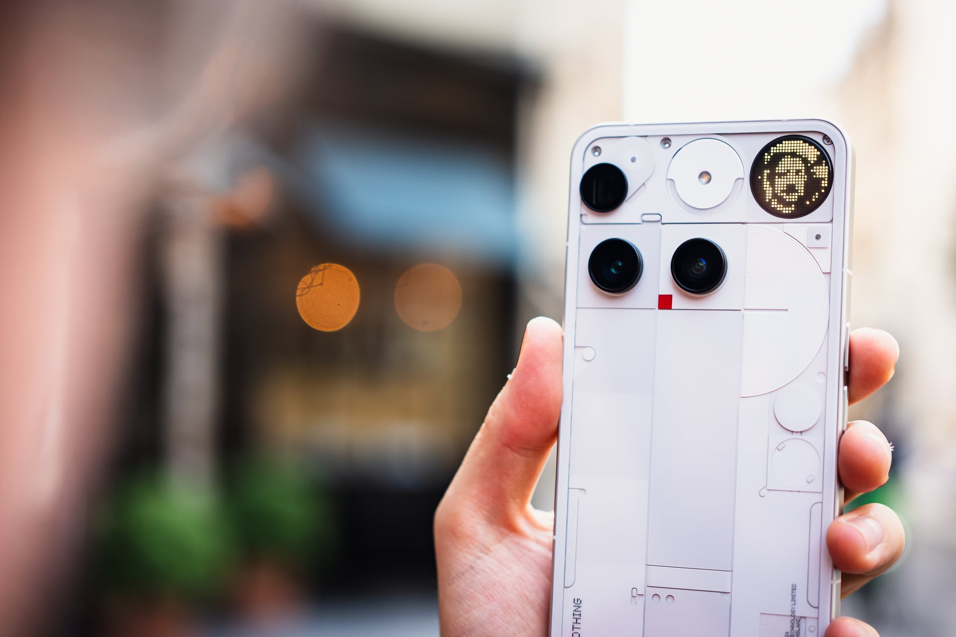 Main tenant un smartphone blanc de dos, révélant trois capteurs photo et un écran circulaire affichant un smiley lumineux