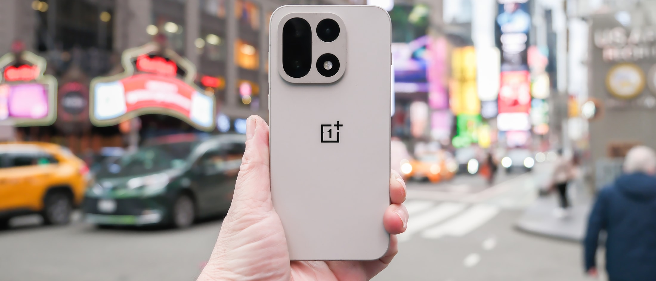 Une main tient un smartphone gris clair arborant le logo OnePlus devant une rue de ville animée et floue avec des taxis jaunes.