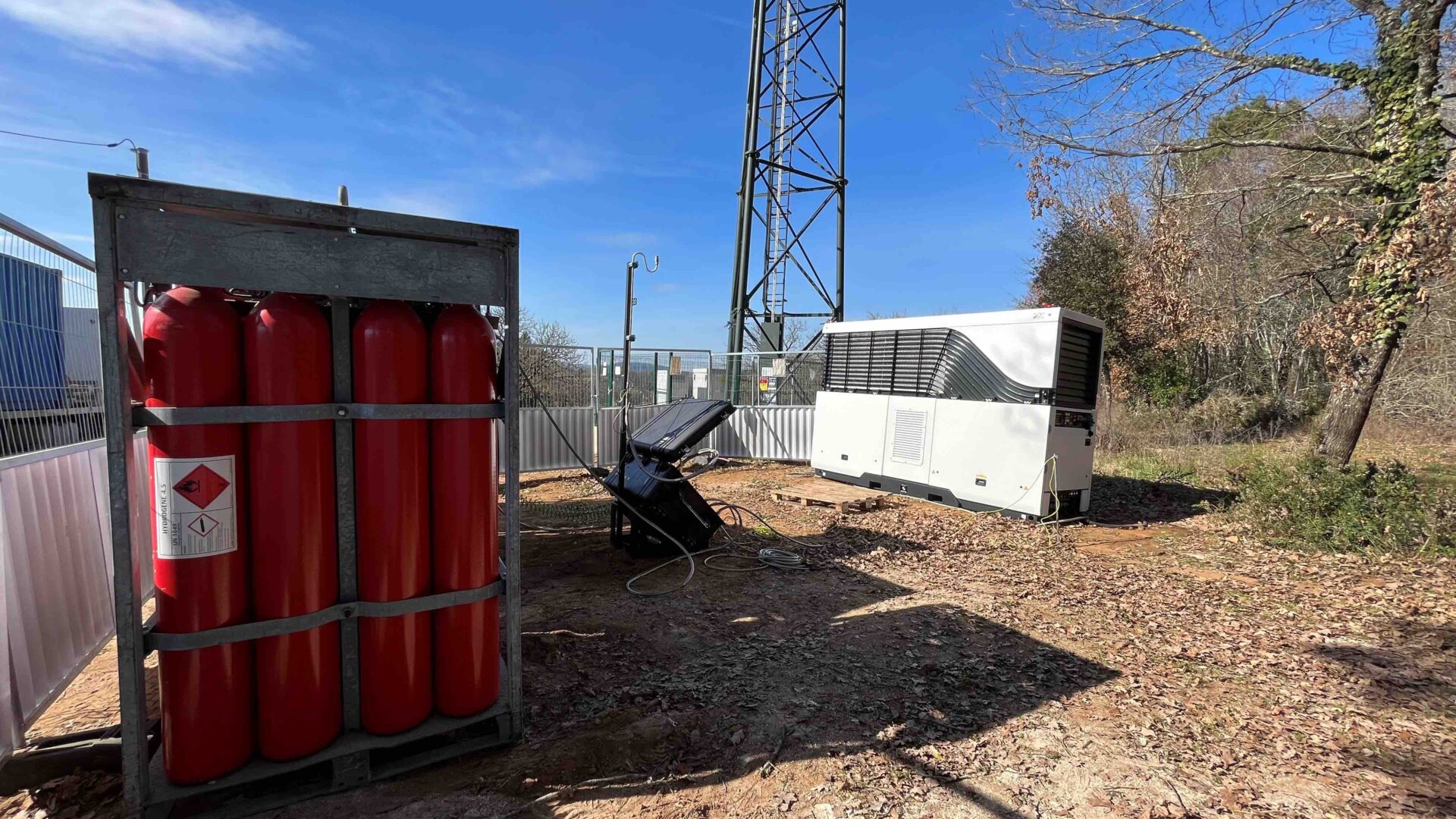 Installation d’antenne-relais avec un pylône métallique, des bonbonnes rouges dans un cadre métallique et un générateur blanc en extérieur.