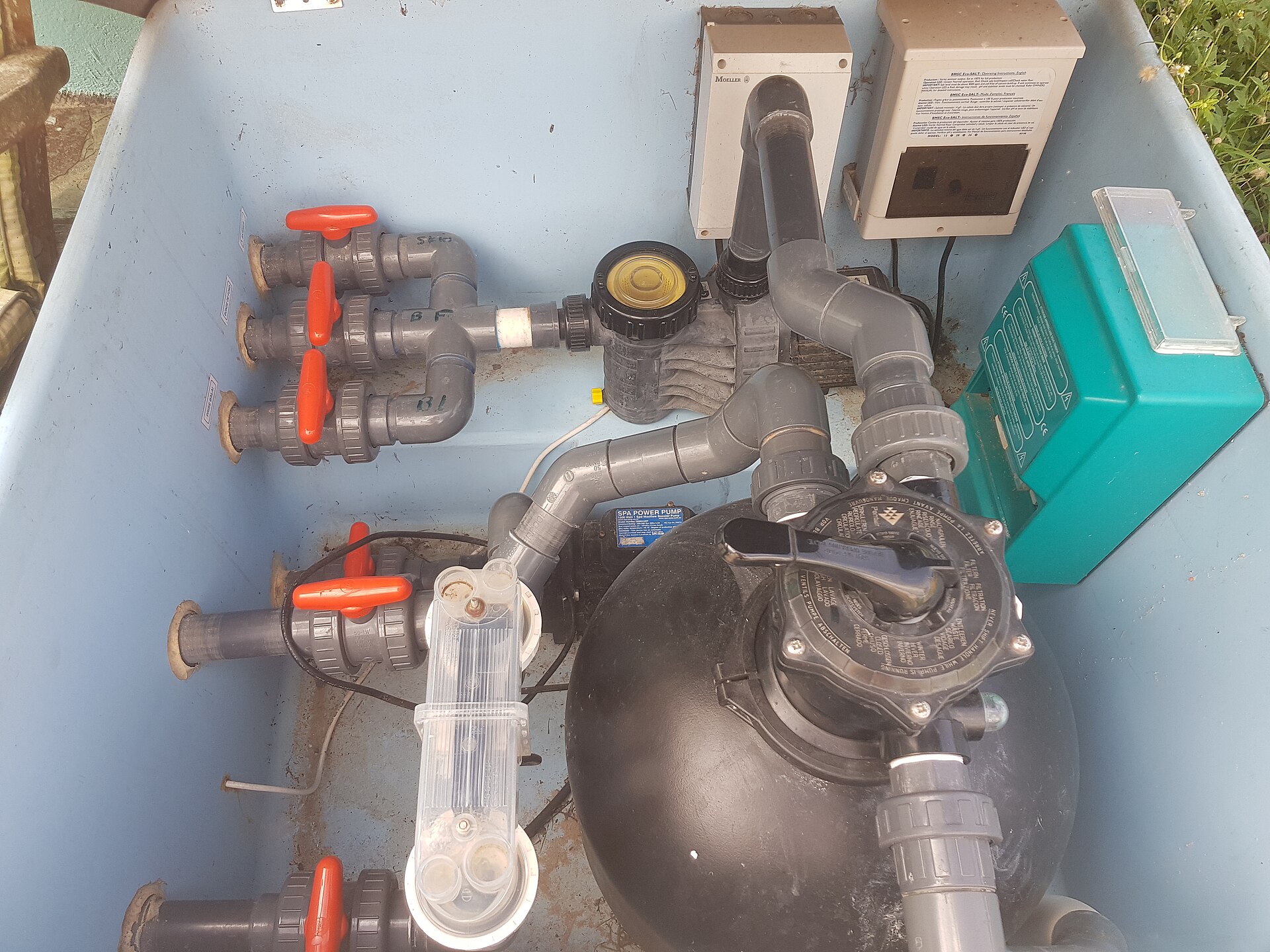 Installation technique de piscine avec tuyauterie grise, vannes rouges, filtre à sable noir et équipements électriques dans un local technique