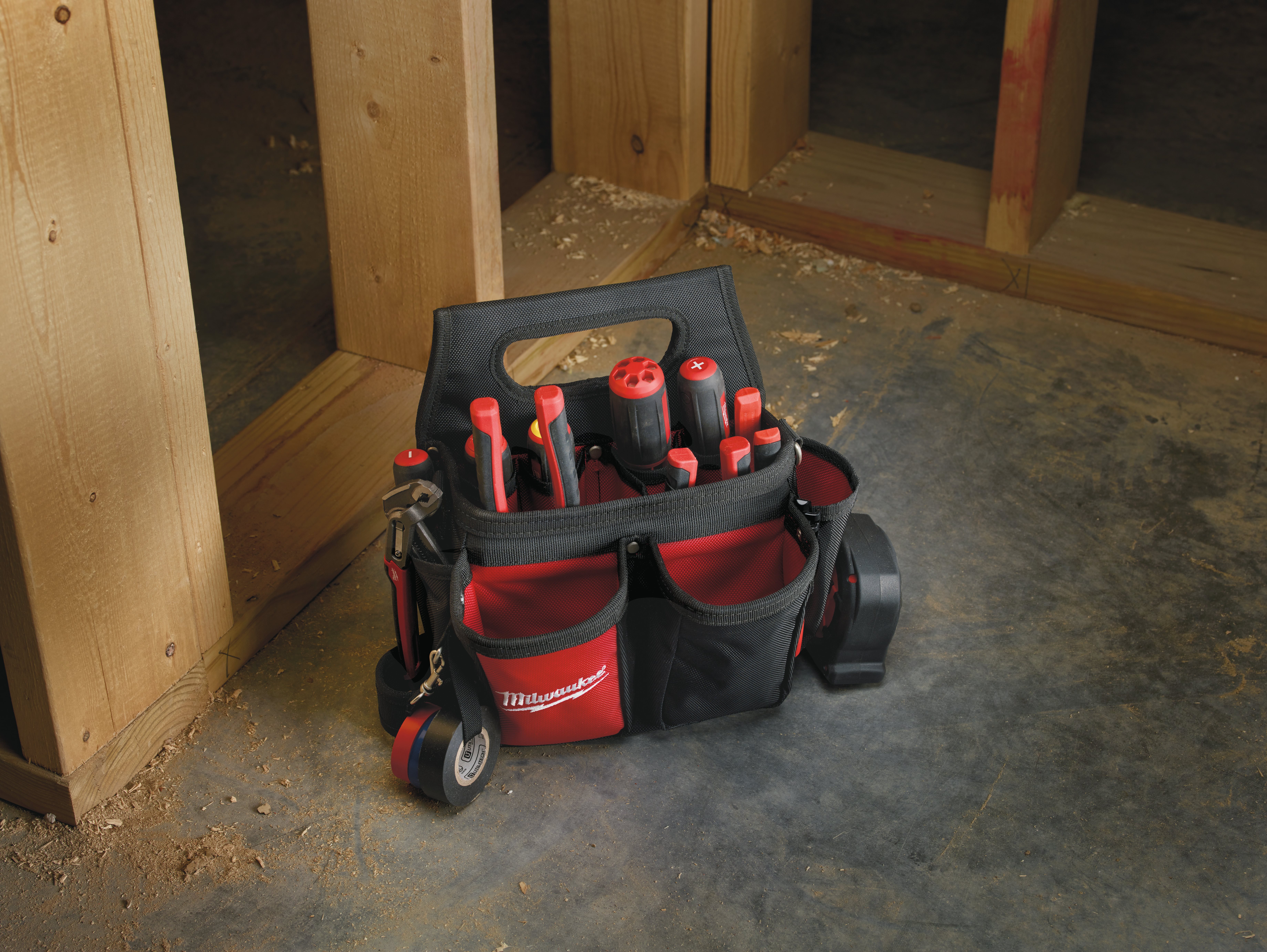 Un sac à outils ouvert noir et rouge de la marque Milwaukee rempli de tournevis et posé au sol dans un chantier en bois
