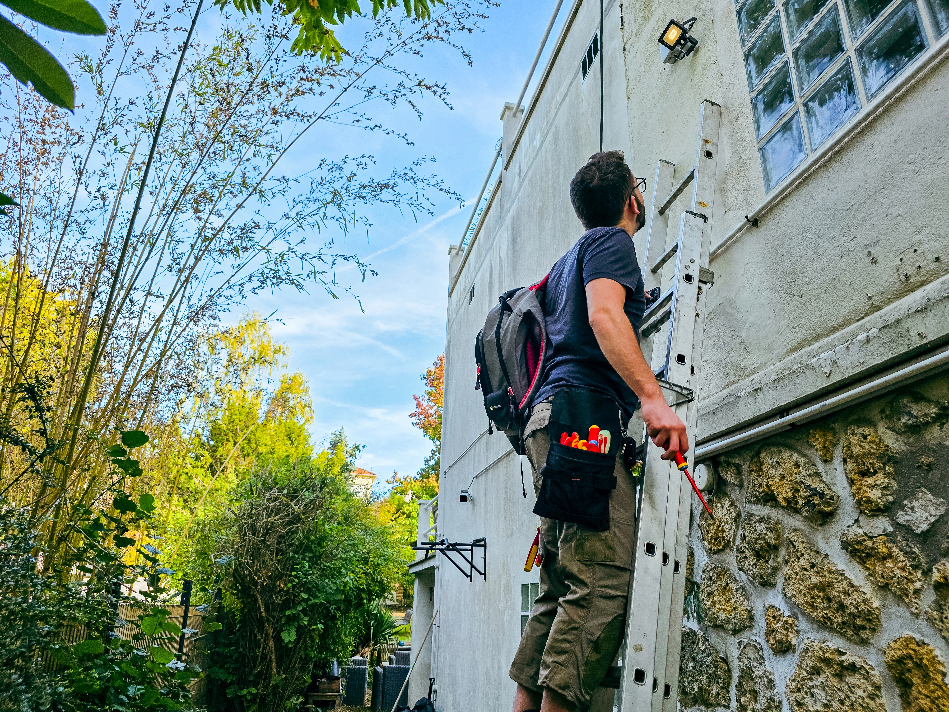 Artisan sur une échelle en façade, portant un sac à dos C.K Magma et tenant un tournevis