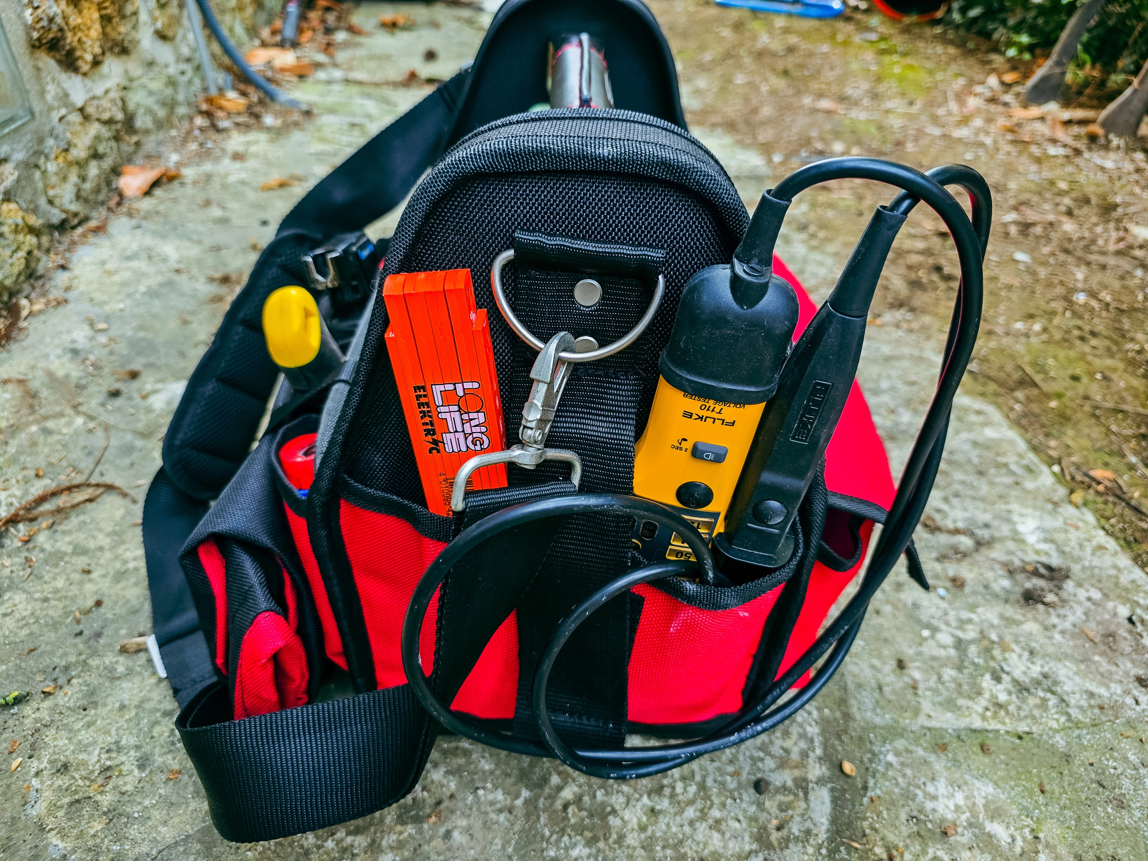 Sac à outils ouvert rouge et noir, vérificateur Fluke T110 et accessoires de mesure répartis dans les poches