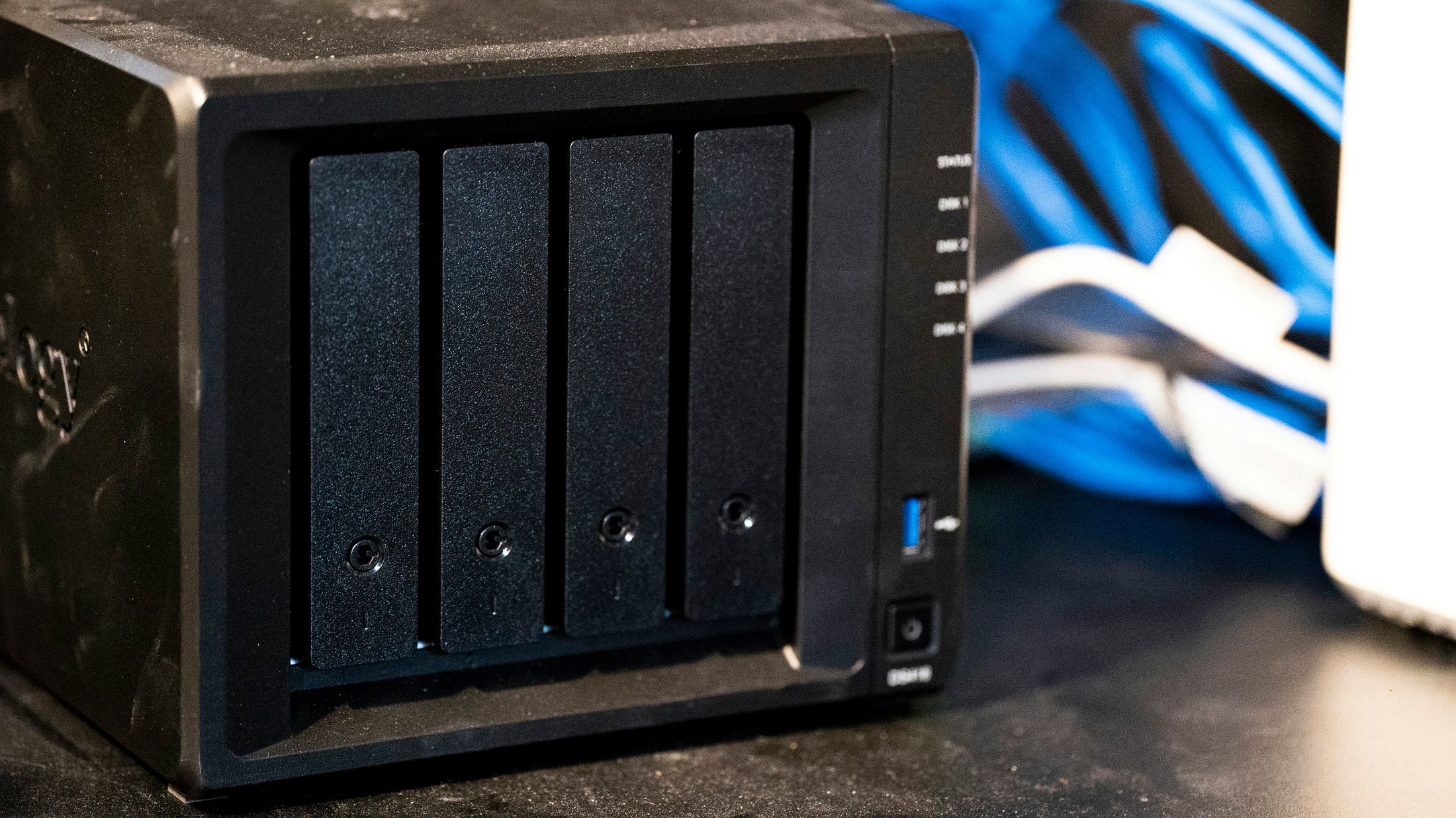 Un gros plan sur la façade d’un boîtier NAS noir à quatre baies de disques durs, avec des voyants d’état et un port USB, posé sur un bureau avec des câbles réseau en arrière-plan.