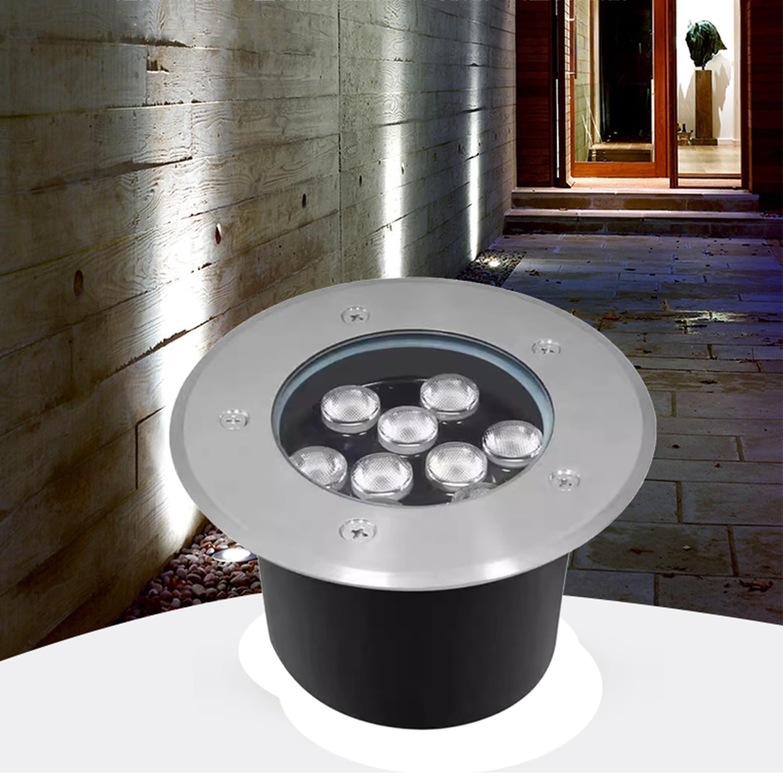 Spot extérieur encastrable rond avec une bordure en acier inoxydable et sept petites lentilles LED, posé sur une surface blanche devant un mur en béton éclairé et une allée pavée.