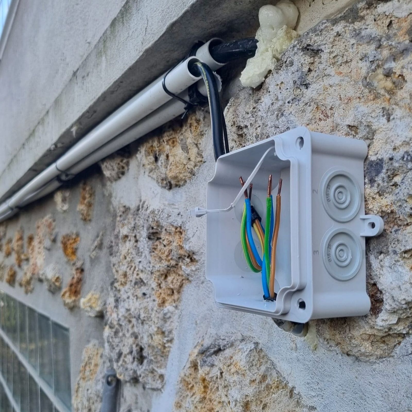 Boîte de dérivation étanche ouverte sur un mur en pierre avec conducteurs rigides dénudés en attente de raccordement