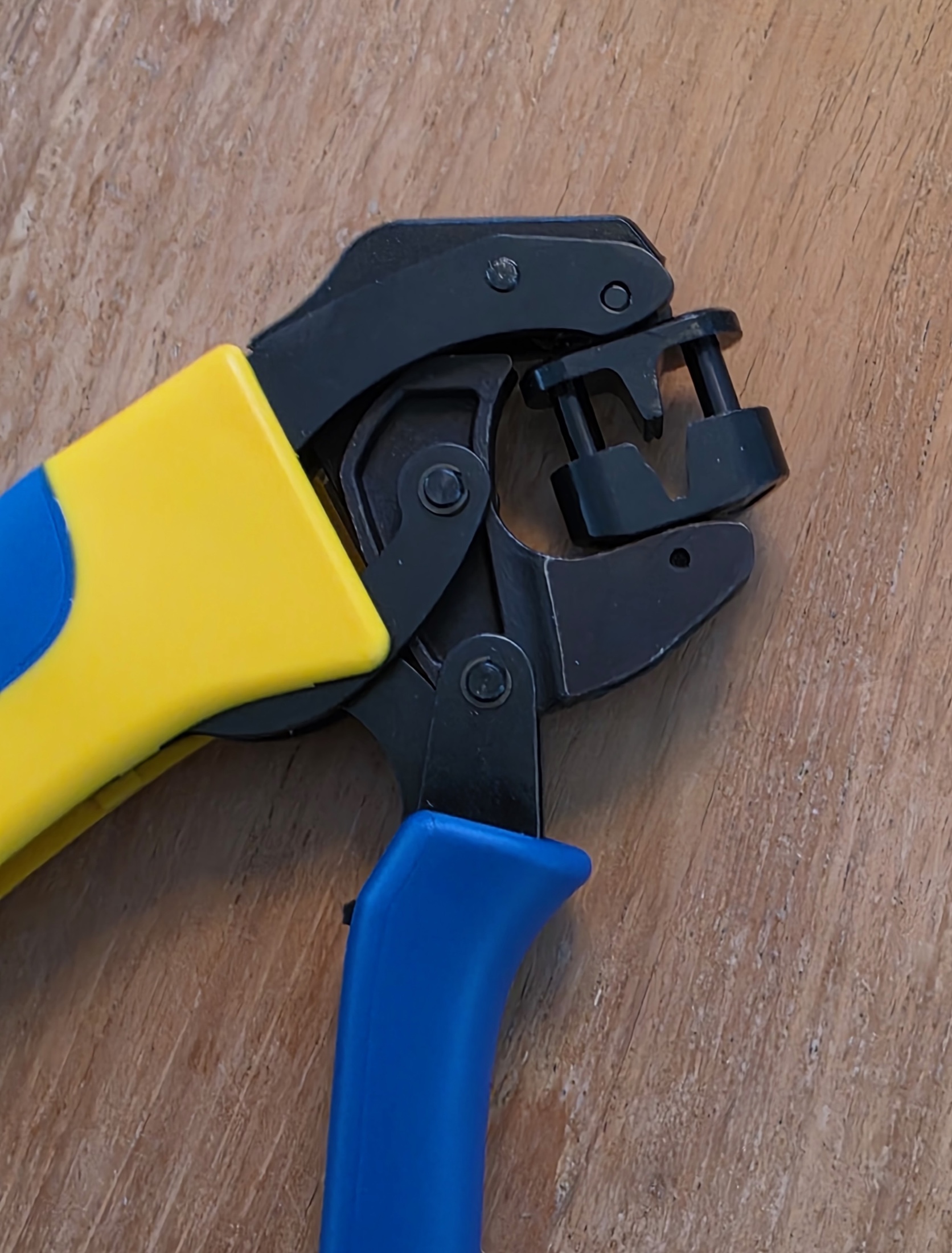 Mâchoires ouvertes d’une pince à sertir noire aux poignées bleues et jaunes, sur une surface en bois